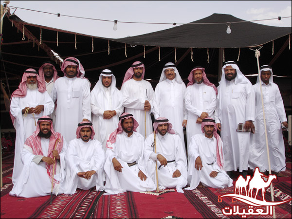 مجموعة من الرجال في زي تقليدي عربي، يتجمعون في موقع خارجي تحت خيمة، مع سجادة حمراء وخلفية من السماء والغيوم.