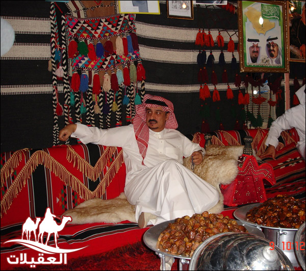 رجل يرتدي الملابس التقليدية السعودية يجلس في مجلس مزين بأقمشة ملونة وأطعمة تقليدية، مثل التمر.