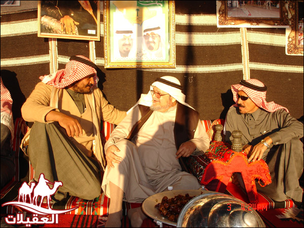 ثلاثة رجال يجلسون معًا في خيمة تقليدية، يتبادلون الحديث. يرتدون الملابس العربية التقليدية ويظهر في الخلفية مجموعة من الصور.