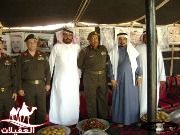 مجموعة من الرجال يرتدون الزي العسكري والتقليدي يقفون معًا داخل خيمة، مع وجود حلويات وأطعمة تقليدية أمامهم وصور على الجدران.