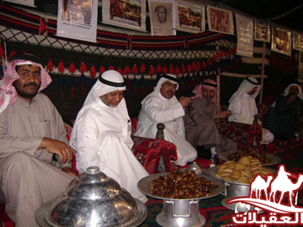 مجموعة من الرجال يرتدون الملابس التقليدية يجلسون في خيمة، مع طعام وأطباق تقليدية حولهم.