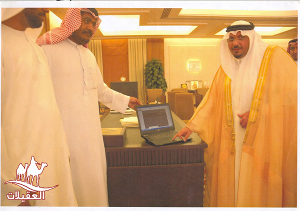 ثلاثة رجال بملابس تقليدية يقفون بجانب طاولة مكتب، يظهرون جهاز لابتوب على الطاولة. الغرفة تحتوي على ديكور مذهل ونباتات.