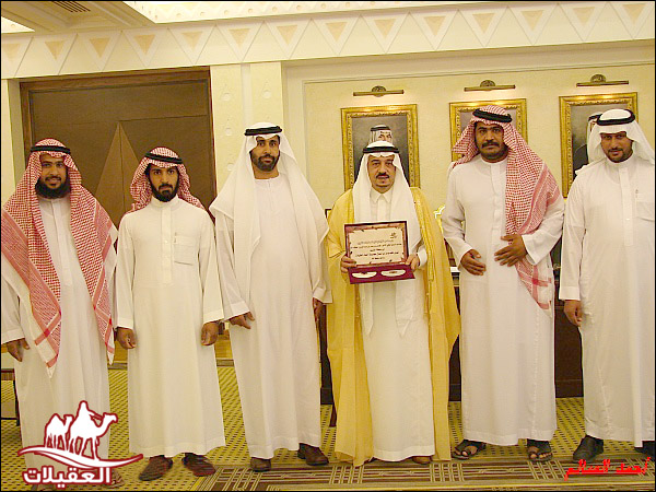 مجموعة من الرجال يرتدون الأزياء التقليدية العربية، يقفون معًا في غرفة داخلية، حيث يحمل أحدهم شهادة أو جائزة.
